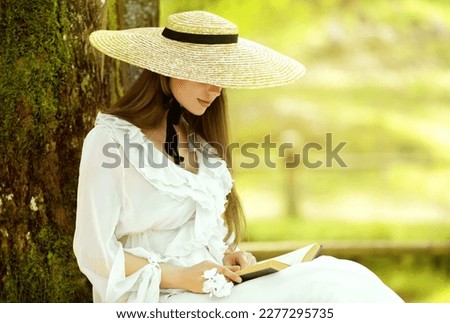 Romantic Girl in White Dress and Straw Sun Hat reading Book sitting under Tree. Young Woman studying in Spring Park. Old Fashioned Elegant Lady outside
