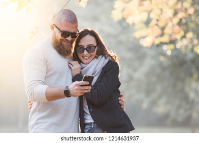 Romantic happy couple having fun on a date. Man with big beard showing something on smartphone to his girlfriend. Autumn sunny sunset mood.