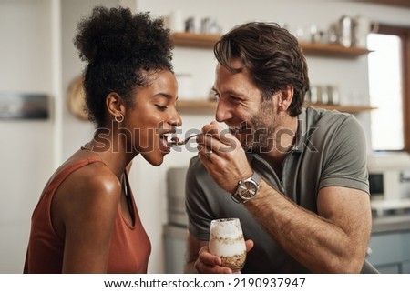 Romantic, happy and interracial couple eating a healthy yogurt together in a cute, sweet and fun kitchen romance. Loving, in love and excited husband feeding his beautiful afro wife delicious