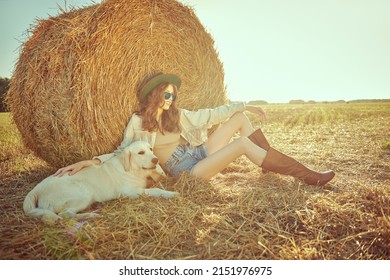 Romantic hippie girl sits by a haystack in a field with her beloved dog. Light from the setting sun. The spirit of adventure and freedom. 