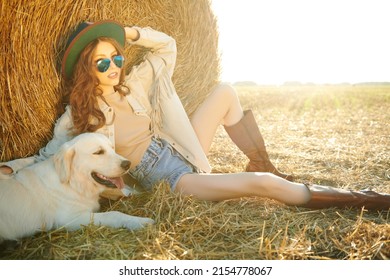 Romantic hippie girl sits by a haystack in a field with her beloved dog. Light from the setting sun. The spirit of adventure and freedom. 