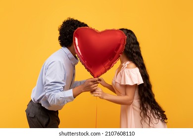 Romantic indian couple kissing behind heart shaped balloon on Valentine's Day, posing over yellow studio background, profile side view. Passionate lovers expressing their affection