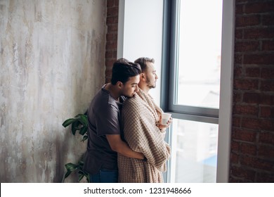 Romantic Indoor Shot Of Caucasian Gay Couple Standing Against Window In Modern Loft Design Studio, Looking At The Street, Dominant Man Taking Care Of His Mate, Tucking Him With Plaid