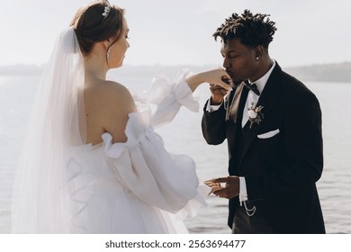 Romantic interracial couple exchanging vows by a serene lake, groom tenderly kissing the bride's hand, celebrating love and unity - Powered by Shutterstock