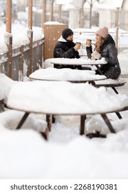 Romantic loving couple walking together on the street in winter, drinking hot drinks on the snow-covered tables of an outdoor cafe. Snow-covered tables of a street cafe. High quality photo - Shutterstock ID 2268190381
