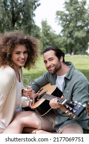 Romantic Man Playing Acoustic Guitar Near Smiling Woman With Glass Of Wine During Picnic