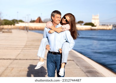 Romantic Mature Couple Having Fun Together While Walking Outdoors On Promenade By The Sea, Cheerful Middle Aged Spouses Enjoying Date Outside, Husband Giving Piggyback Ride To Cheerful Wife - Powered by Shutterstock