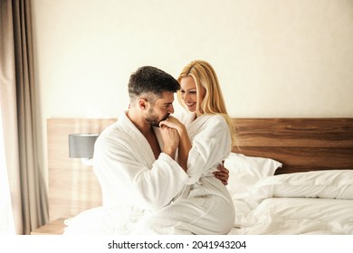 Romantic mornings at the hotel. A man and a woman in white robes and on the white sheets of a warm hotel room cuddling in bed. A kiss in a woman's feminine hand, macho man. Love, smiling, honeymoon - Shutterstock ID 2041943204