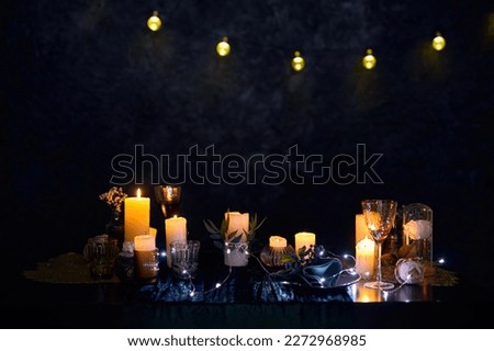 A romantic night scene in a dark restaurant with a table set with decorations, glowing candles, and glasses of red wine, illuminated by a string of ice lights