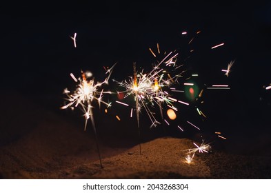 Romantic Nighttime Background With Sparks. Beach Sand At Night With Sparklers. Sparks Of Bright Bengal Lights Burning On The Beach. Defocus, Blur, Noise, Grain Effect.
