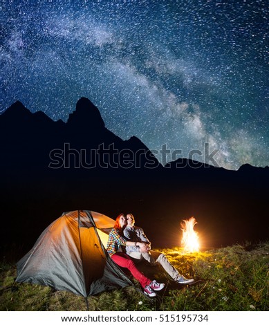 Romantic pair lovers looking to the shines starry sky and Milky way in the camping at night near campfire. On the background silhouette of the high mountains. Low light