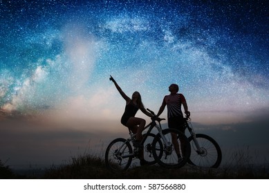 Romantic Pair With Mountain Bikes On The Hill Under Night Starry Sky. Woman Shows Man At The Stars. Bottom View. Night Landscape With Colorful Milky Way.