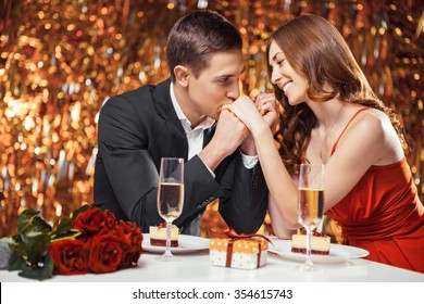 Romantic photo of beautiful couple on glitter gold background. Couple having date at Valentine's Day. Lovers having dinner. There are glasses with champagne, desserts, roses and gift on table - Powered by Shutterstock