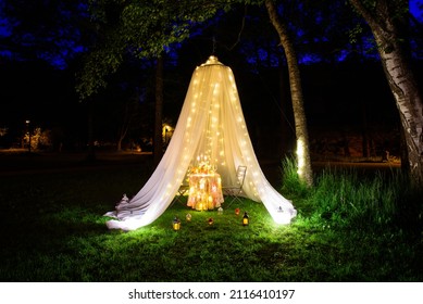 A romantic place for a date or marriage proposal in a park on the lake, in a beautiful tent and decorated with candles and garlands. A table set with fruit and a bottle of wine. - Shutterstock ID 2116410197