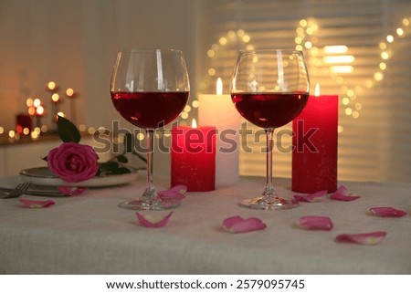 Romantic place setting with red wine, burning candles, rose and petals on table against blurred lights