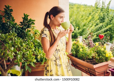 Romantic portrait of beautiful young woman wearing yellow dress, relaxing on the balcony between many green plants, holding cup of tea or coffee