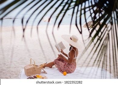 Romantic portrait of  stylish asian woman sit on beach carpet blanket having picnic with basket of fruits, wearing top, long skirts and big large straw hat, holding reading book. - Powered by Shutterstock