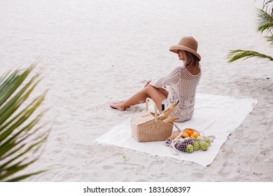 Romantic portrait of stylish woman sit on beach carpet blanket having picnic with basket of fruits on white sand beach, wearing knitted stylish clothes and straw hat