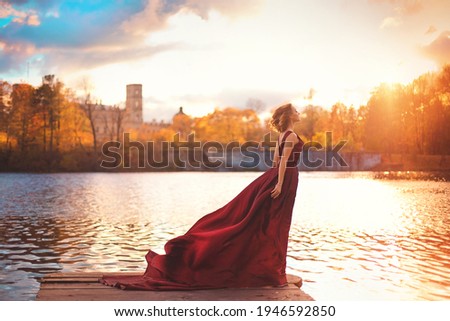 Romantic portrait of a young girl in a long red dress standing near the columns of an old manor house on the background of autumn nature