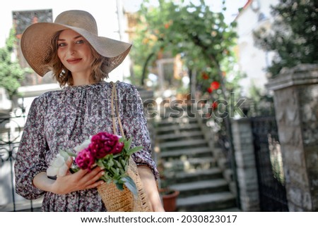 Romantic portrait of young woman dressed style dress and straw hat, femal hold peonies against the garden of the house