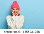 Romantic positive young European woman smiles gently, has white perfect teeth, touches both cheeks, has friendly look, wears pink hat with pompon and white sweater, models against blue wall.