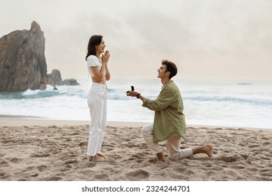 Romantic proposal on the seashore. Loving young man with engagement ring making proposal to happy woman on beach on coastline, side view - Powered by Shutterstock