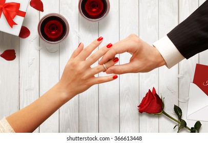 Romantic Proposal, Wedding Or Valentine's Day Scene. Man Putting Engagement Ring. Background With Glasses Of Wine, Gift, Rose, Envelope On Wooden Table.