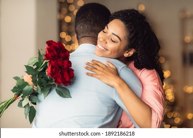 Romantic Relationship. Glad smiling african american woman hugging her man and holding bouquet of red roses. Girlfriend with bunch of flowers embracing boyfriend, celebrating anniversary