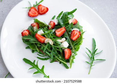 Romantic salad of strawberry with mix of herbs, arugula, spinach leaves, feta cheese and cashew nuts in heart shape on white plate. Romantic meal for St Valentine Day celebration.