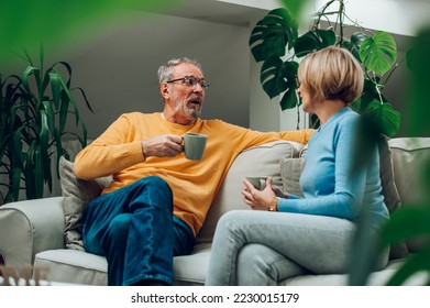 Romantic senior couple laughing ant talking to each other while sitting on sofa at home and drinking coffee. Affectionate elderly married couple enjoying sweet tender cozy moment. Focus on a man. - Powered by Shutterstock