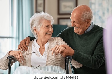 Romantic senior husband embracing disabled wife sitting on wheelchair at nursing home. Cheerful old couple embracing and looking at each other with love. Elderly man holding hands of handicapped woman