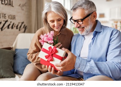 Romantic senior man congratulating happy wife on Valentines Day or March 8 at home, surprised elderly female receiving gift box with red bow and tulips bouquet from loving husband. Selective focus - Powered by Shutterstock