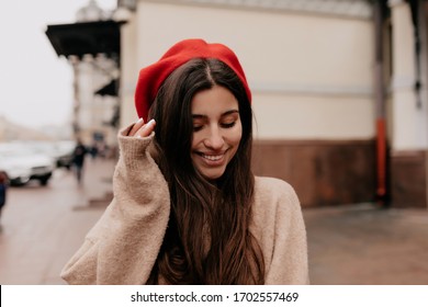 Romantic shy woman with long dark hair wearing red cap and beige jacket walking down the street on background of old city and smiling with closed eyes in spring day