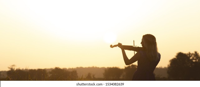 Romantic Silhouette Of Young Woman With A Violin At Dawn On River Bank, Elegant Girl Playing A Musical Instrument On Nature, Concept Music And Inspiration