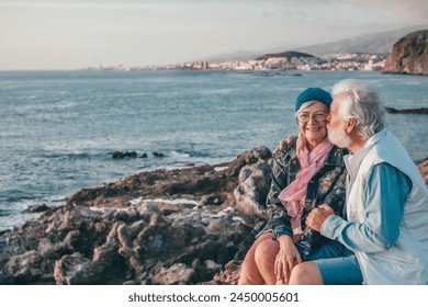 Romantic smiling senior couple sitting on the rocky beach at sea enjoying vacation and retirement at sunset light. Relaxed couple of retirees exchange kisses and cuddles - Powered by Shutterstock