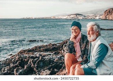 Romantic smiling senior couple sitting on the rocky beach at sea enjoying vacation and retirement at sunset light. Relaxed couple of retirees looking at horizon over water - Powered by Shutterstock