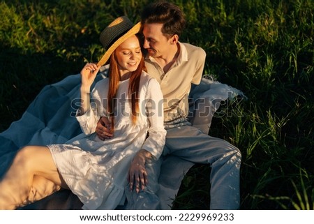 Romantic smiling young couple in love with closed eyes sitting hugging together on beautiful green meadow in summer evening during golden sunset, contemplating and daydreaming their future.