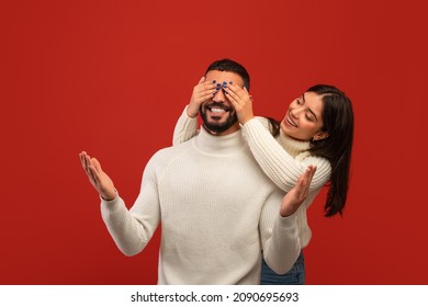 Romantic Surprise Concept. Cheerful Arab Woman Covering Her Boyfriend Eyes From Back, Standing Behind Him, Couple Posing On Red Studio Background