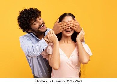 Romantic Surprise Concept. Portrait Of Happy Indian Guy Covering His Pretty Girlfriend Eyes From Back, Standing Behind Her, Couple Posing On Yellow Studio Background, Banner