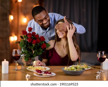 Romantic Surprise. Happy black man covering white girlfriend's eyes and giving red roses bouquet. Loving interracial couple having dinner in restaurant, celebrating Valentine's day together - Powered by Shutterstock