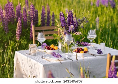 Romantic table decor for a loving couple on the blooming meadow with purple lupines. Two glasses of wine, flowers in a vase, silverware, fruits, wooden furniture and picnic basket. Sunset, golden hour - Powered by Shutterstock