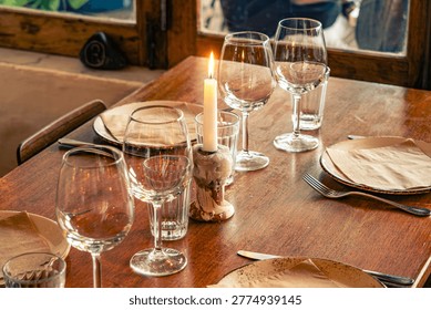 Romantic table setting with burning candle and wine glasses in a rustic restaurant.
A close-up, warm-toned photograph of a set wooden dining table inside a cozy restaurant. The focus is on a single lit white candle in a rustic ceramic holder, creating a soft glow. The table is arranged with elegant empty wine glasses, water tumblers, ceramic plates, and craft paper napkins. The background shows a blurred window and wooden interior, evoking an intimate atmosphere for a dinner date or a social gathering.