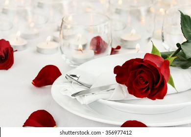 Romantic table setting with long stem red rose and candles burning in the background. Shallow depth of field with selective focus on rose.
