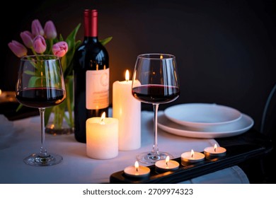 A romantic table setting with two glasses of red wine, a bottle of wine, lit candles, and a bouquet of pink tulips.