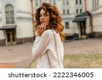 Romantic tender woman with curly hair wearing white blouse looking around at camera while walking in the city in warm sunny evening. Portrait of cheerful pretty woman looking away and smiling. 
