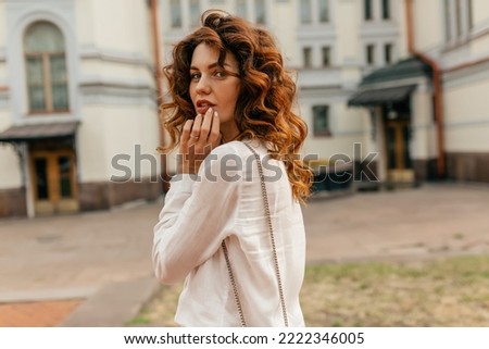 Romantic tender woman with curly hair wearing white blouse looking around at camera while walking in the city in warm sunny evening. Portrait of cheerful pretty woman looking away and smiling. 