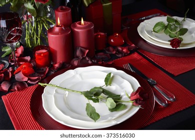 Romantic Valentine Candle Light Dinner For Two Table Setting For Two With Red Roses, Gift And Burning Candles Against A Black Background. 