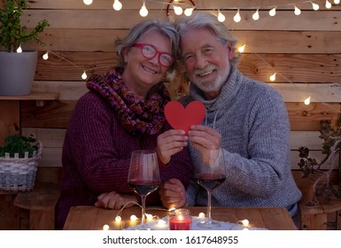 A Romantic Valentine's Day For An Elderly Couple Sitting Outdoors On A Rustic Wooden Bench Holding A Red Heart In Their Hands. Two Older People Celebrate Love With Wine Glasses And Sparkling Lights