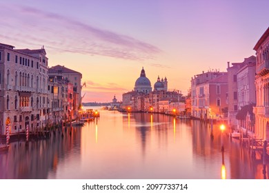 Romantic Venice at dawn, sunrise. Cityscape image of Grand Canal in Venice, with Santa Maria della Salute Basilica reflected in calm sea. Street lights reflected in calm water.