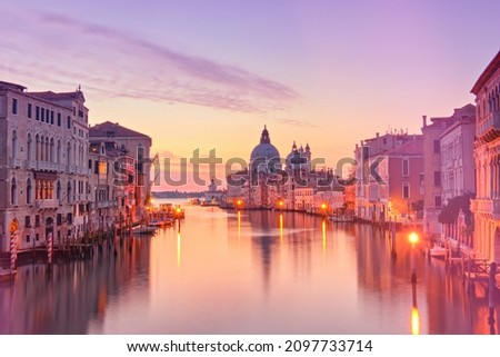 Romantic Venice at dawn, sunrise. Cityscape image of Grand Canal in Venice, with Santa Maria della Salute Basilica reflected in calm sea. Street lights reflected in calm water.